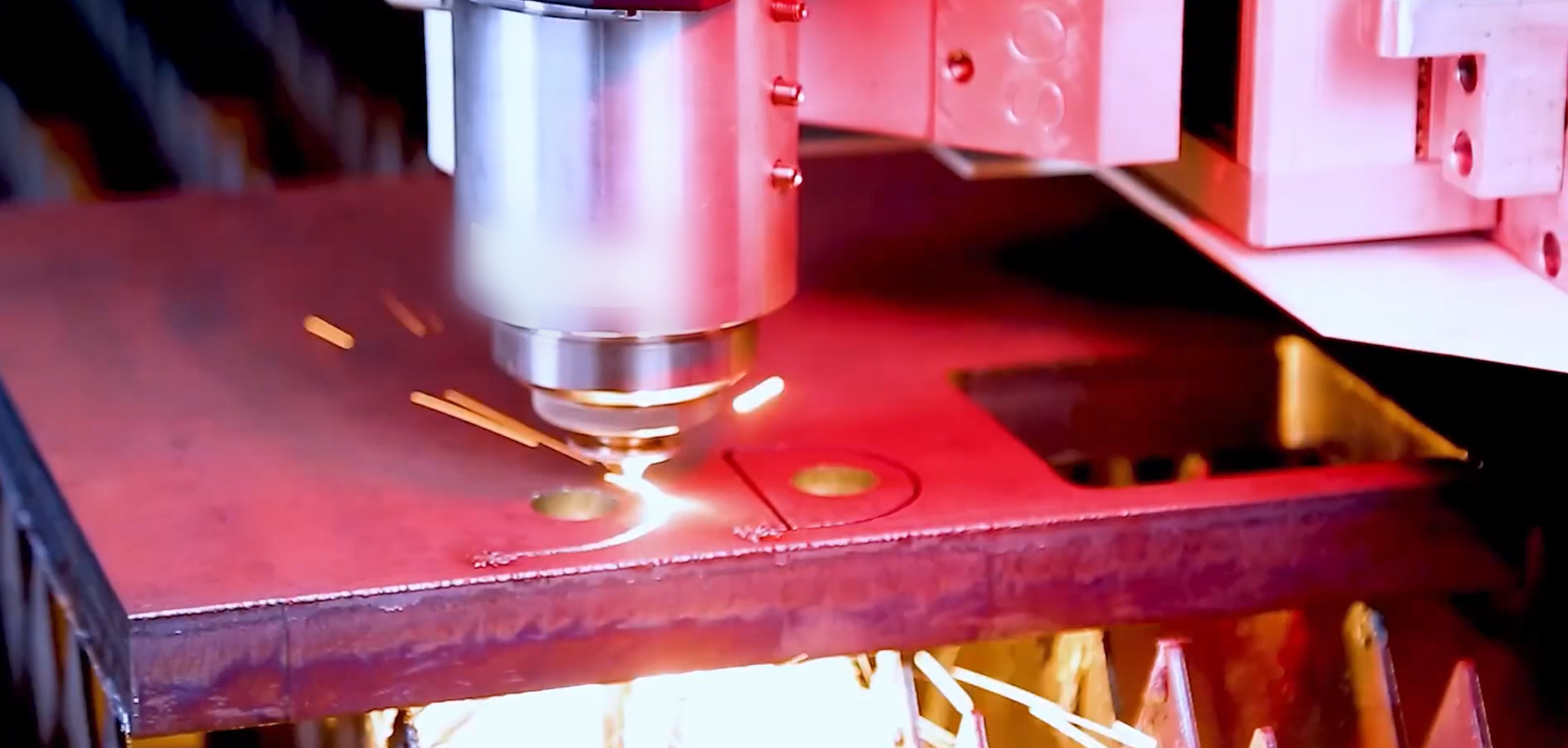 A fiber laser cutting head works through steel plate on a slat bed, finished holes visible in the sheet and molten light flaring under the cut.